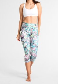 Kobieta w białym staniku sportowym i kolorowych legginsach w tropikalny wzór, stojąca boso na jednolitym tle.