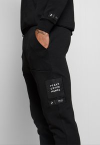 Pantalon de survêtement noir en tissu doux, avec poches avant, un écusson portant le texte "PEAK PERFORMANCE" et une petite étiquette sur le côté.