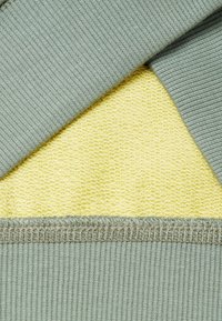 Baumwollmischgewebe mit einem gerippten grünen Außenbereich und einem strukturierten gelben Innenbereich, mit kontrastierenden Nähten entlang der Kanten.