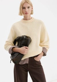 Kremowy, oversize'owy sweter z dzianiny z prążkowanym dekoltem, w połączeniu z brązową koszulą w paski i ciemnobrązowymi spodniami, trzymający czarną skórzaną torbę.