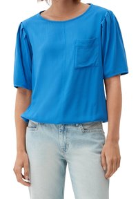 Frau in einer leuchtend blauen kurzärmligen Bluse mit Brusttasche und hell gewaschenen Jeans vor einem weißen Hintergrund.