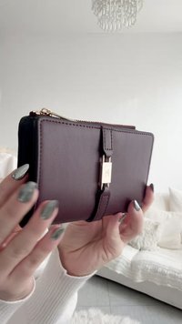 Handen met nagels in groen marmerpatroon houden een donkerbruin leren portemonnee vast met een gouden sluiting en ritssluiting in een lichte, minimalistische kamer.