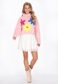 Rosa gestrickter Pullover mit hohem Kragen, verziert mit großen Blumenapplikationen in Rosa, Gelb und Blau. Trägt einen Rock mit weißen Spitzenbesätzen.