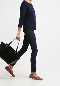 Námořnický svetr s ribovaným vzorem, tmavé úzké džíny, hnědé mokasíny a velká černá kabelka. Oblečení představuje ležérní, moderní design.