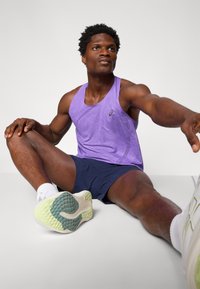 Lila Sporttanktop mit strukturiertem Design, marineblaue Shorts und weiße Laufschuhe mit einer grünen und türkisfarbenen Sohle. Sitzende Position.
