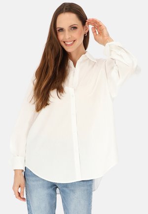 Uśmiechnięta kobieta o długich brązowych włosach nosząca luźną białą koszulę zapinaną na guziki i jasnoniebieskie dżinsy, podnosząca rękę przy głowie na białym tle.