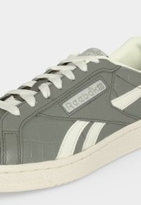 Γκρι αθλητικό παπούτσι Reebok με λευκά κορδόνια, λευκές πλευρικές ρίγες, υφή στη σόλα και λογότυπο στην πλευρική επιφάνεια και στη γλώσσα.