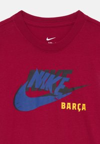 Rdeča bombažna majica s kratkimi rokavi, na kateri je velik, zbledel črno-modri Nike logo z besedo "BARÇA" v poudarjeni rumeni barvi. Okrugel izrez.