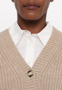 Šviesiai ruda mezginė kardiganas su gilia V formos iškirpte, su tekstūruotomis raukšlėmis ir dideliu apvaliu rudu mygtuku virš baltos marškinių apykaklės.