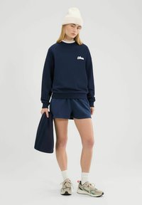 Joven mujer vestida con sudadera azul marino y pantalones cortos, gorro blanco, calcetas blancas y zapatillas beige, sosteniendo una bolsa de tela azul marino.