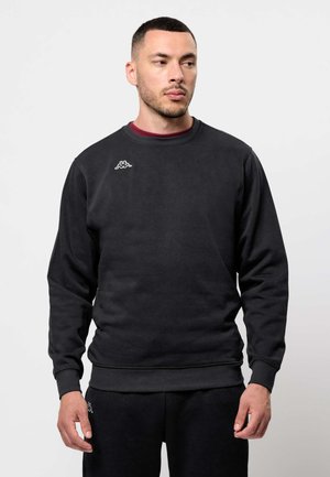 Férfi fekete pulóvert és hozzáillő fekete nadrágot visel, melyeken kis fehér logó látható a mellkason és a combon, egyszerű háttér előtt áll.