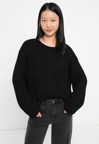 Giovane donna con capelli neri lisci che indossa un maglione nero morbido e jeans grigio scuro, in piedi con le mani nelle tasche su uno sfondo bianco.