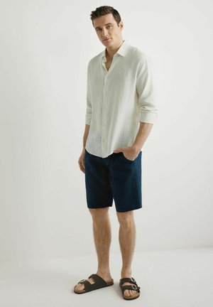 Młody mężczyzna stoi z jedną ręką w kieszeni, ubrany w białą, długorękawową koszulę na guziki, granatowe szorty i czarne sandały na sprzączkę na jednolitym tle.