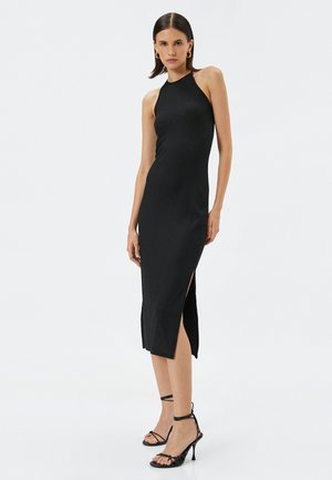Vestido negro sin mangas con una silueta ajustada, textura acanalada y una gran abertura en el lado izquierdo. Llevado con tacones negros con tiras.