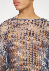 Gros plan d'une personne portant un haut en maille lâche multicolore bleu et blanc, laissant entrevoir la peau nue en dessous.