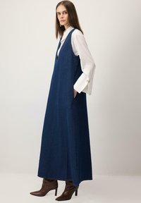 Tumši zilas džinsa kombinezona kleita ar dziļu V veida izgriezumu, sānu kabatām un vaļēju siluetu. Valkāta pār baltu kreklu un brūnās papēžu zābakos.