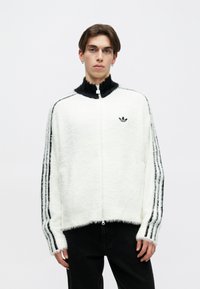 Chaqueta blanca de felpa con cremallera y cuello negro acentuado, que presenta tres franjas negras en las mangas y un pequeño logo negro en el pecho.