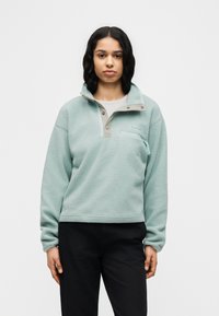 Νεαρή γυναίκα με σκούρα σγουρά μαλλιά, ντυμένη με ανοιχτό μπλε φλις πουλόβερ και μαύρο παντελόνι, στέκεται μπροστά σε ένα απλό λευκό φόντο.