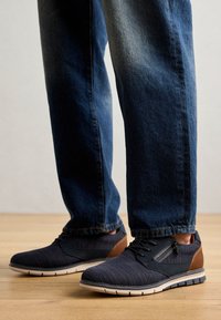 Baskets légères de couleur bleu foncé avec un tissu texturé, des accents marron et une semelle rembourrée, associées à un jean en denim bleu sur un sol en bois.