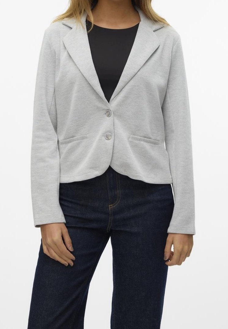 Femme portant un blazer gris clair boutonné sur un haut noir et un jean bleu foncé, debout, les mains relâchées sur les côtés.
