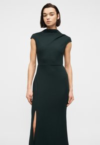 Νέα γυναίκα με αμάνικο σκούρο πράσινο φόρεμα με ψηλό λαιμόκοψη και σκίσιμο στο μπούτι, να στέκεται μπροστά σε απλό φωτεινό φόντο.