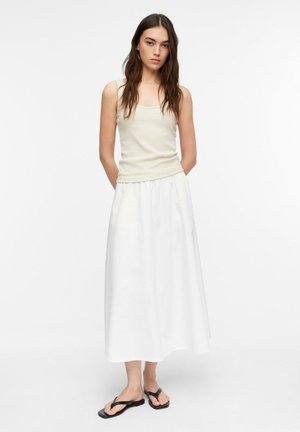 Kobieta o długich ciemnych włosach, ubrana w kremowy top bez rękawów, białą spódnicę midi i czarne płaskie sandały, stojąca na białym tle.