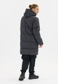 Manteau long rembourré gris foncé avec une capuche, présentant un design matelassé et des poignets élastiques, associé à des joggings noirs et des chaussures de sport noires.