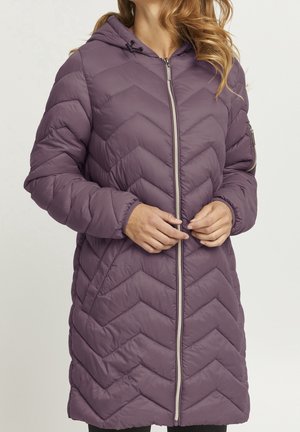 Femme portant une longue veste matelassée à capuche violette, les mains serrant la fermeture éclair frontale, sur un fond clair uni.