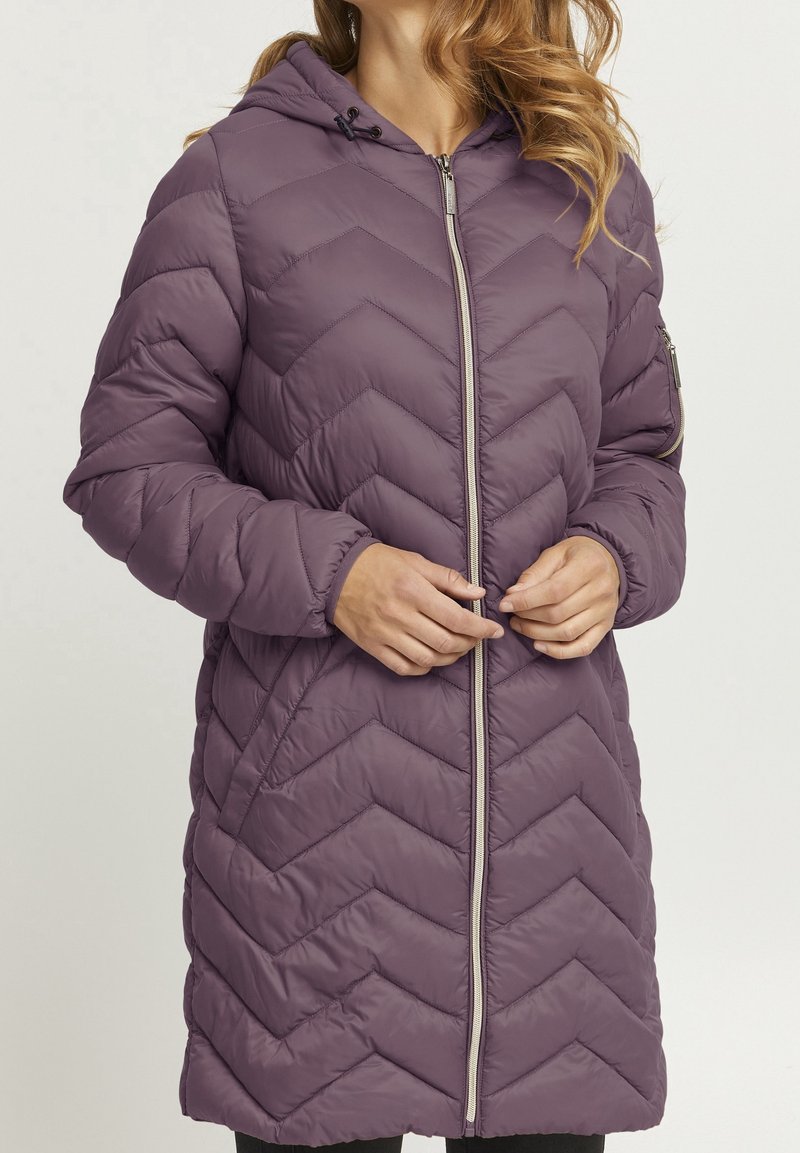 Femme portant une longue veste matelassée à capuche violette, les mains serrant la fermeture éclair frontale, sur un fond clair uni.