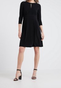 Musta mekko, jossa on kolme neljännes-sleevit, pyöreä pääntie ja avainreikätiedot. Liukas kangas, istuva vyötärö ja kellotettu helma. Mustat hihna-korkokengät.