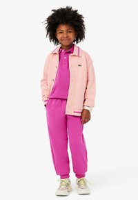 Růžová bomber bunda se pruhovanými manžetami, fialové polo tričko a ladící růžové tepláky. Tenisky mají žluté tkaničky a šedé akcenty.