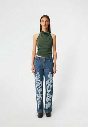 Kvinde iført en ærmeløs grøn halterneck-top og blå jeans med stor hvid blomsterbroderi står foran en ensfarvet baggrund.