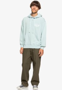 Light blue hoodie made of soft cotton, featuring a front pouch pocket and white graphic on the left side. Paired with loose olive green pants.