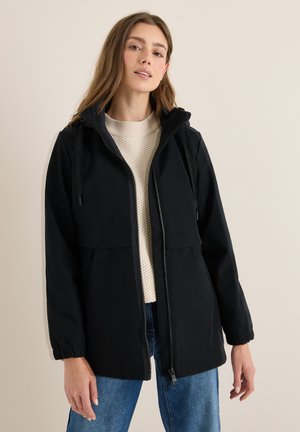 Veste noire imperméable avec fermeture éclair avant, capuche avec cordon de serrage et poignets élastiques, superposée sur un pull léger et un jean bleu.