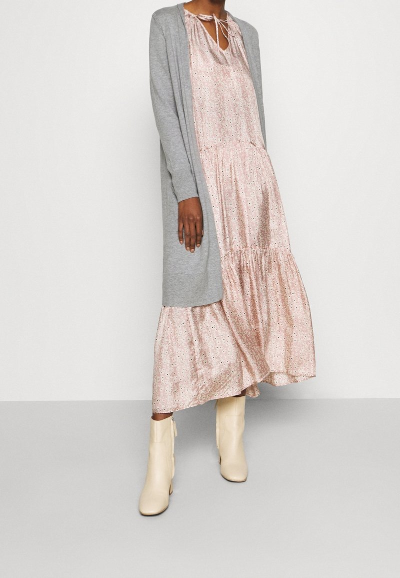 Grauer langer Cardigan über einem rosa Blumen Kleid. Das Kleid hat einen Bindekragen und einen gestuften Rüschensaum. Kombiniert mit cremefarbenen Ankle Boots.