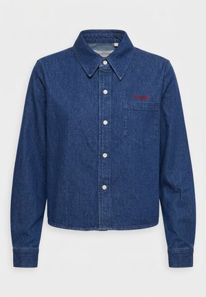 Pessoa vestindo uma camisa de ganga azul escura abotoada com bolso no peito e calças brancas largas, de pé com uma mão atrás das costas.
