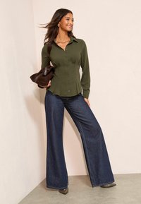 Cămașă verde măsliniu cu nasturi și pliuri, asortată cu blugi evazați din denim închis la culoare. Modelul ține o geantă maro închis și poartă pantofi cu toc.