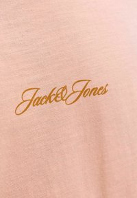Ροζ ύφασμα με χρυσό κίτρινο λογότυπο "Jack & Jones" με καλλιγραφική γραφή τυπωμένο στην επιφάνεια, που δείχνει την υφή του υφάσματος.