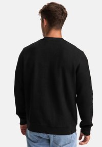 Maglione nero in tessuto testurizzato con un motivo geometrico, collo a giro e polsini a coste. Indossato con pantaloni in denim chiaro; vista posteriore.