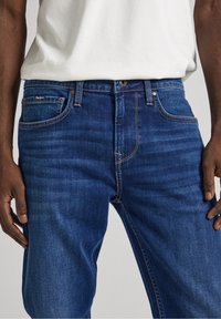 Mörkblå denimjeans med kontrasterande orange sömmar, femfickorsdesign och subtil branding på bakfickan. Slät textur.