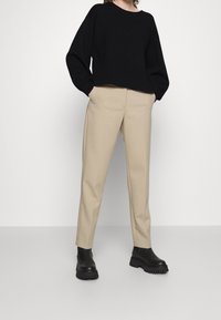 Pull en maille noir, pantalon beige ajusté et bottines noires à semelles épaisses. Le modèle se tient contre un fond blanc.