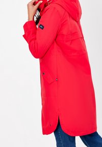 Impermeable rojo con capucha, mangas largas y un dobladillo ligeramente curvado. Cuenta con cierre frontal de botones y un pequeño parche con logo en la manga.