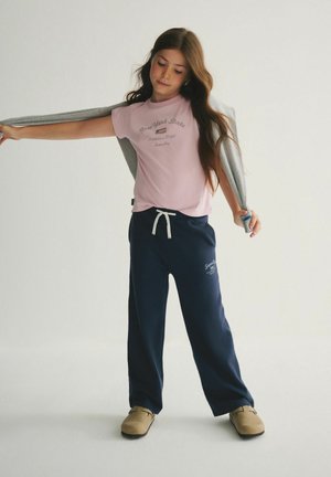 Tüdruk reguleerimas halli jopet sirutatud kätega, kannab roosat "New York State" T-särki, tumesiniseid pükse ja beeže kingi ühetaustalisel heledal taustal.