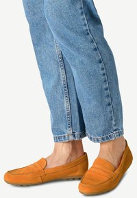 Oranžové semišové loaferky se švem na přední straně, plochou podrážkou a minimalistickým designem. Komplementovány světle modrými, mírně zkrácenými džíny.