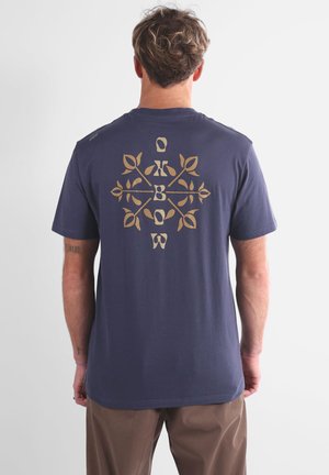T-shirt bleu marine à manches courtes en coton, avec un design graphique doré symétrique dans le dos, orné d'un motif floral et du texte de la marque.