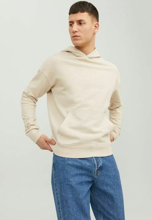 Beigefarbener Hoodie mit einer vorderen Tasche, verstellbarem Kordelzug und gerippten Bündchen. Kombiniert mit hellblauen Denim-Jeans.
