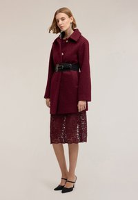 Burgundský texturovaný kabát s velkými knoflíky, se zavazováním v pase, přes burgundskou krajkovou sukni. Černé lodičky s špičatou špičkou doplňují outfit.