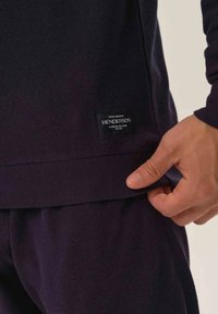 Ciemnofioletowy materiał bluzy o fakturze waflowej. Na dolnym brzegu znajduje się czarna metka z napisem "HENDERSON", a ręka dostosowuje pas.