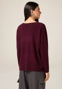 Maglione in maglia bordeaux con vestibilità comoda e spalle scese, caratterizzato da una scollatura ampia e orlo a coste, abbinato a pantaloni grigi.