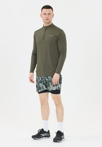 Olivgrünes Langarmhemd mit einem halben Reißverschluss, kombiniert mit gemusterten Shorts in Grau- und Grüntönen, sowie schwarzen Sportschuhen.